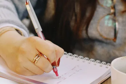 Close-up of a woman's hand writing notes in a notebook with a cup of coffee nearby.