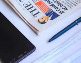 Flat lay of a newspaper, face mask, smartphone, and pen on a white background, capturing modern essentials.