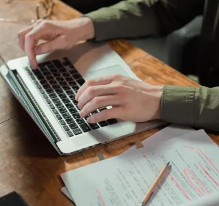 Focused person typing on laptop with notes and papers beside on wooden table.
