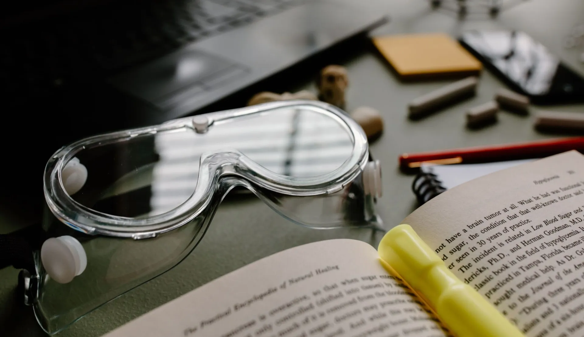 Close-up of lab goggles with an open book and study materials in a science lab setting.