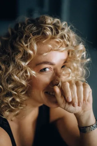 Close-up portrait of a cheerful woman with curly blonde hair covering her mouth playfully.