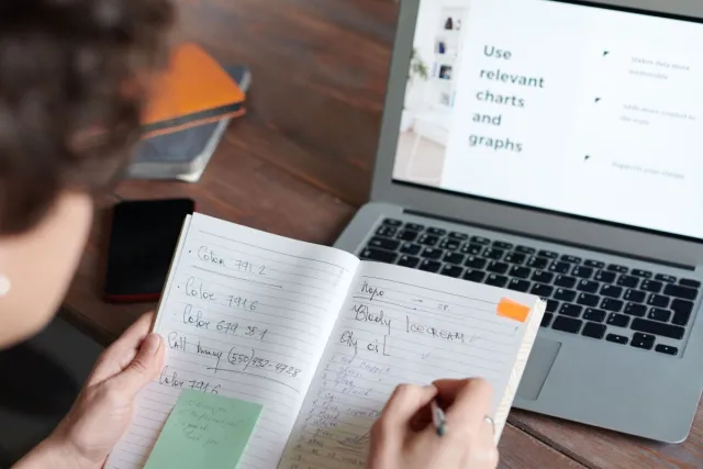 Individual writing notes at a laptop with a digital chart presentation indoors.