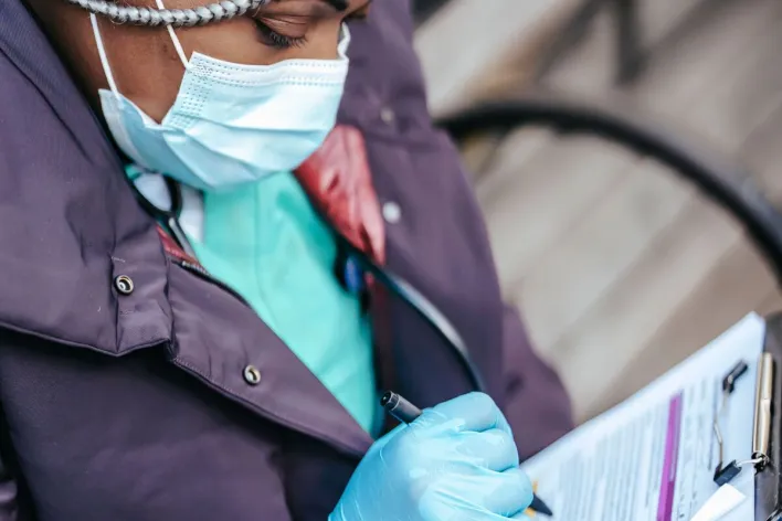Adult black woman nurse in medical uniform and protective mask with gloves and stethoscope sitting on bench while writing information in papers in clipboard
