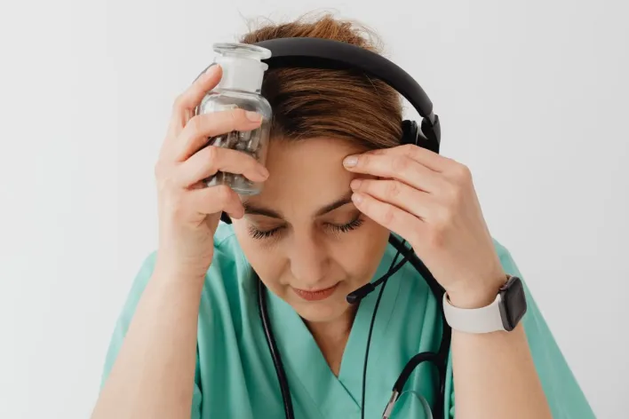 A stressed female medical professional holds a jar of medicine, wearing headphones and a stethoscope.