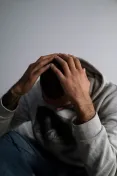 A man in a hoodie sits indoors, appearing contemplative or sad, with a focus on his hands and posture.