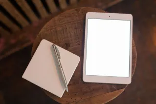Overhead view of a tablet with blank screen and notebook with pen on wooden table.