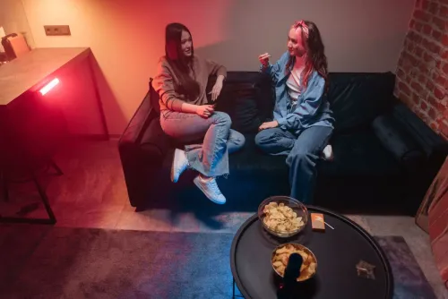Two women relaxing on a couch indoors, illuminated by warm red lighting, sharing a casual conversation.