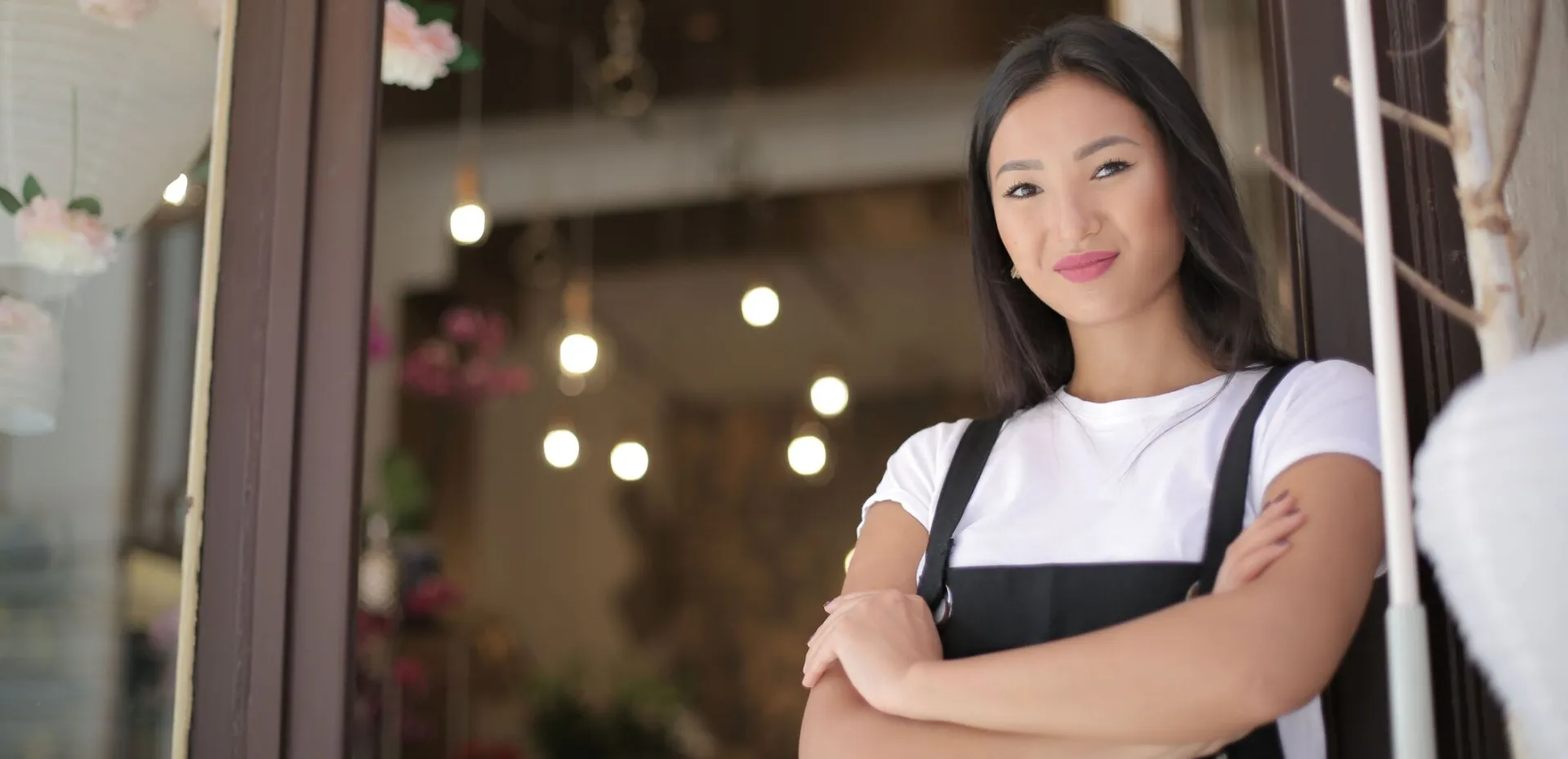 From below of young confident Asian female standing with crossed arms at entrance of floral shop and smiling at camera