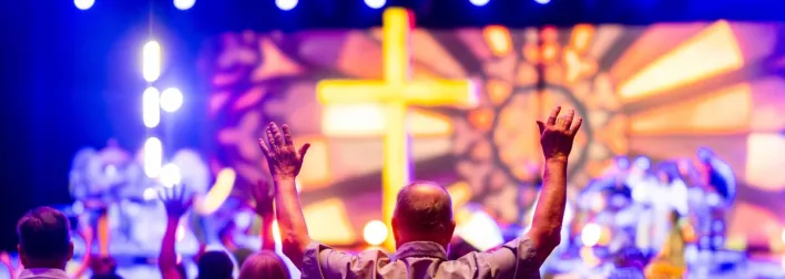 A lively worship service with people praising, colorful lights, and a cross stage backdrop.