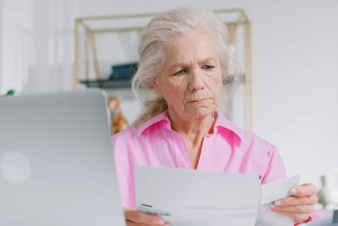 Elderly woman in pink blouse reviewing documents at home office desk.