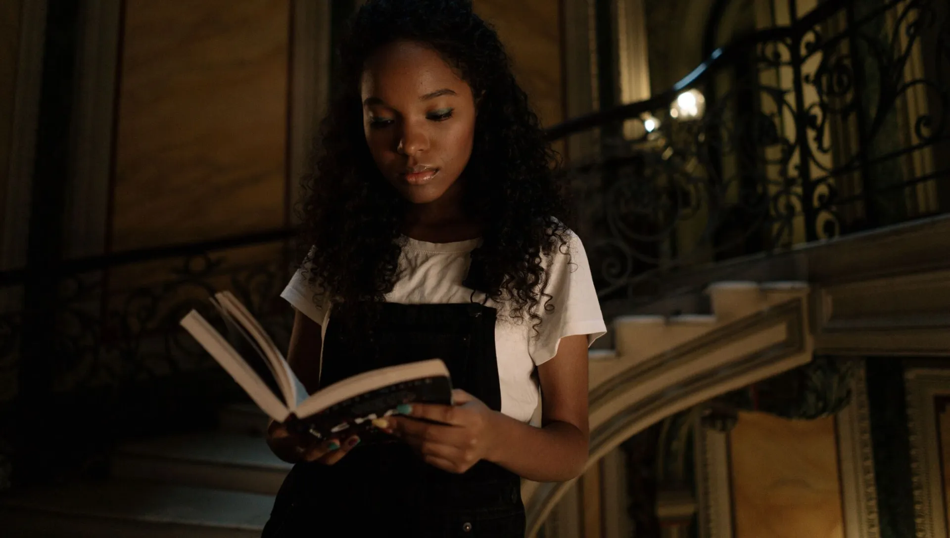 A young woman with curly hair reads a book while standing on an ornate staircase, immersed in thought.