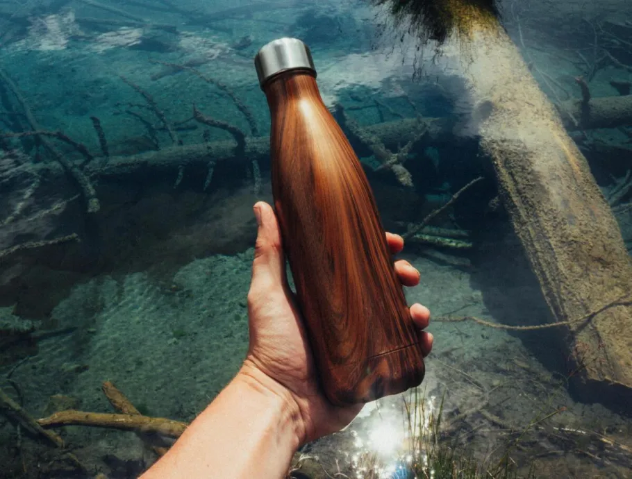 Close-up of a hand holding a wooden water bottle over a clear stream, showcasing eco-friendly lifestyle.