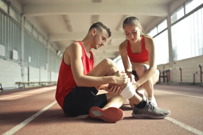 Side view of athlete sitting on rubber track in sports hall while sportive female partner helping to band leg after injury