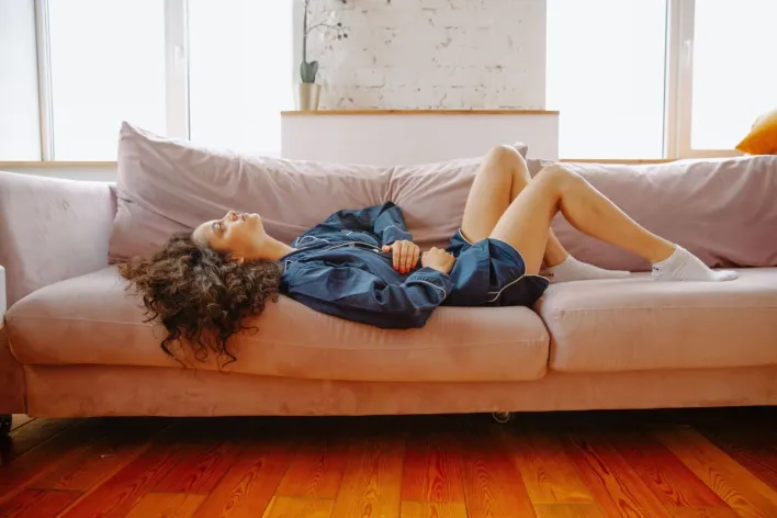 A woman in casual wear experiences abdominal discomfort while lying on a pink couch indoors.