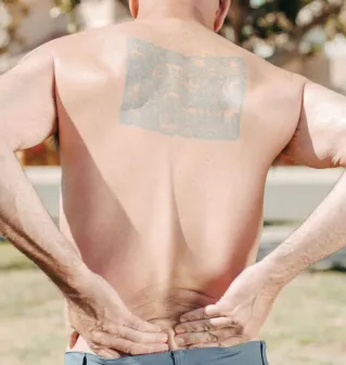 Shirtless bald man from behind holding his lower back, outdoors in sunny weather, with a large tattoo.