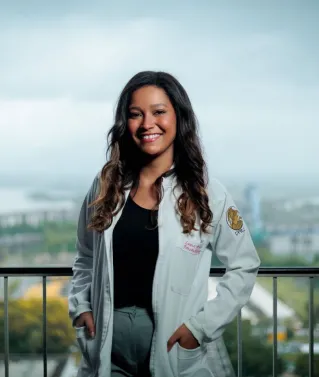 Smiling female doctor in white coat posing on a balcony with a cityscape background.