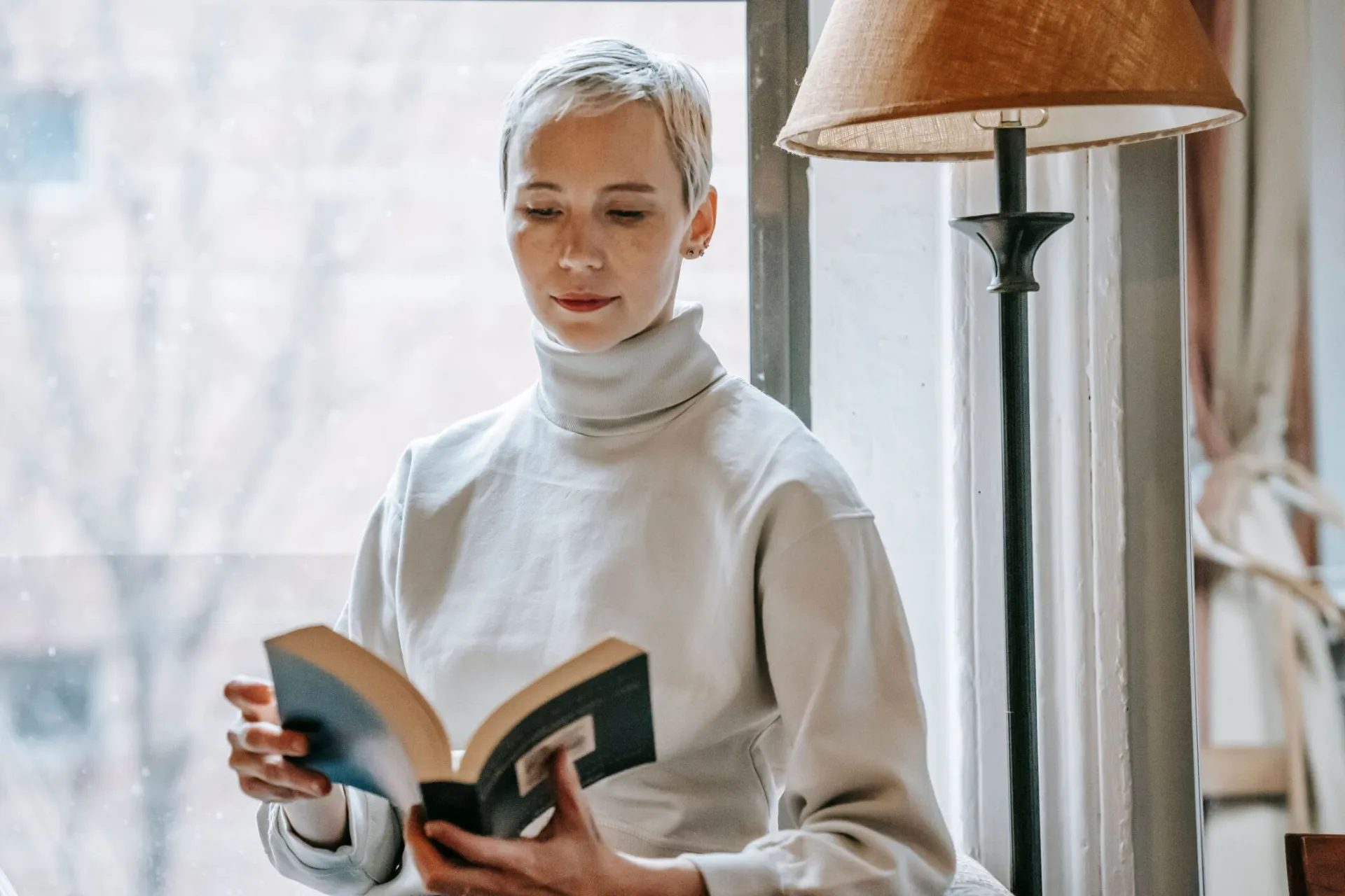Focused smart female with short hair reading interesting book near lamp and window in daytime