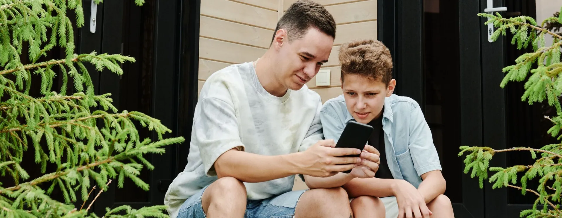 A father and son sitting on porch steps, bonding over a smartphone in a modern outdoor setting.