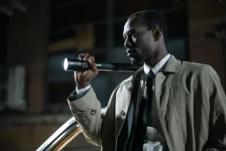 African-American detective in a beige trench coat holding a flashlight outdoors at night.