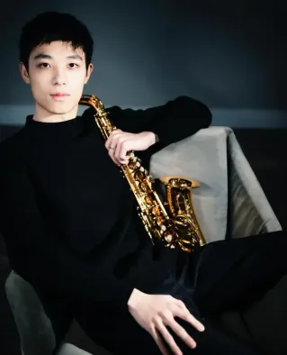 A young male musician singing into a microphone against a dark indoor background.