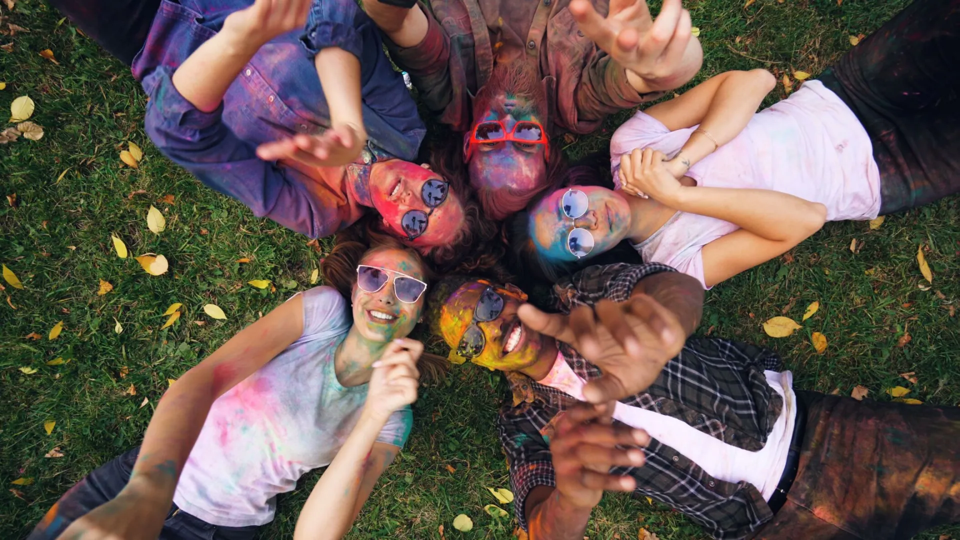 A group of young people covered in colorful powder celebrate Holi outdoors, lying on grass.