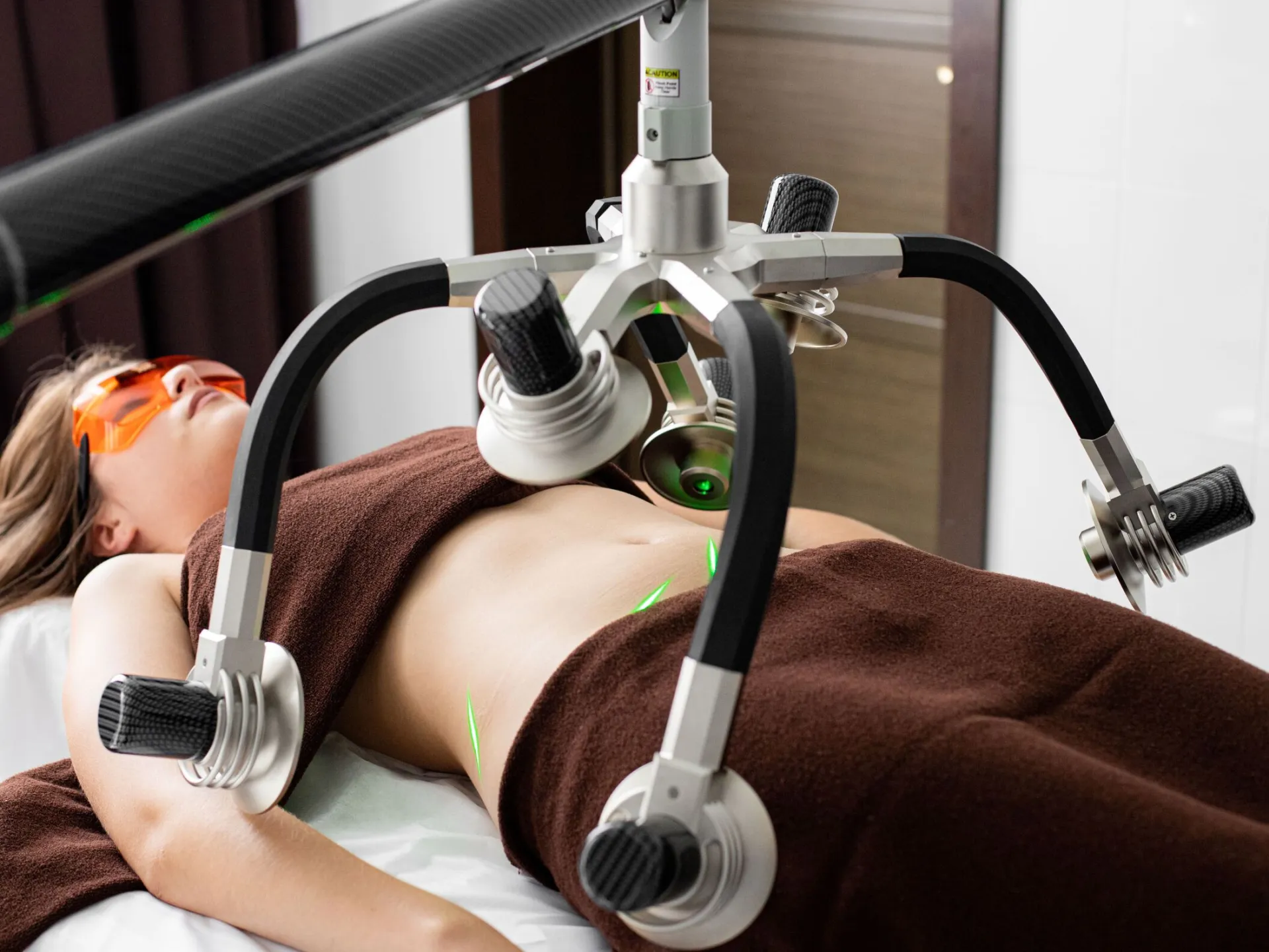 Female client lying on table under lasers of modern weight loss machine on belly during liposuction procedure in medical clinic