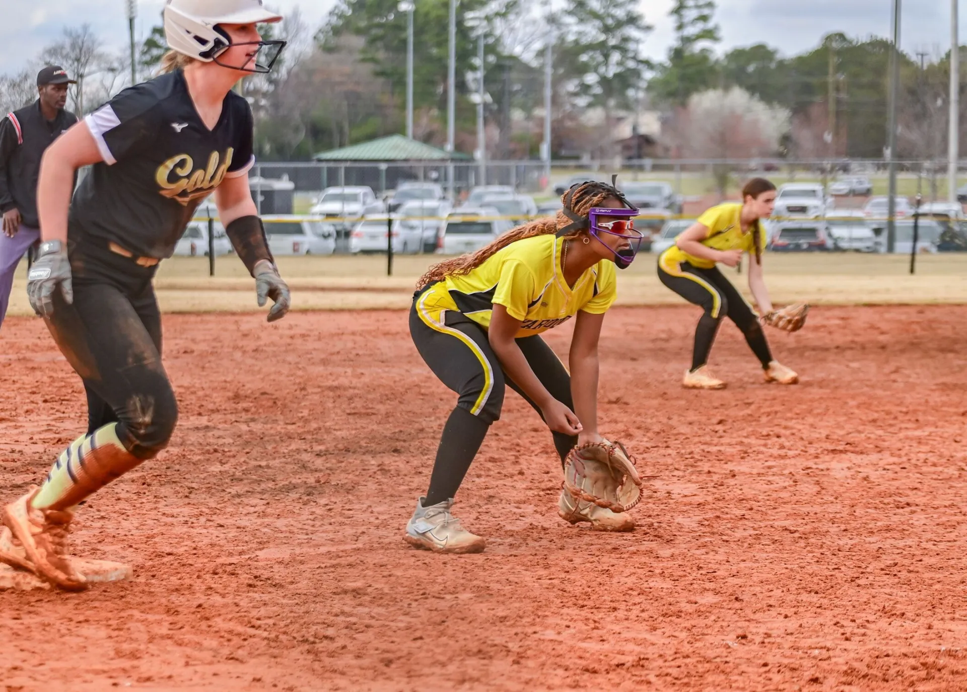 Dynamic shot of female softball players in mid-game action on an outdoor red clay field.