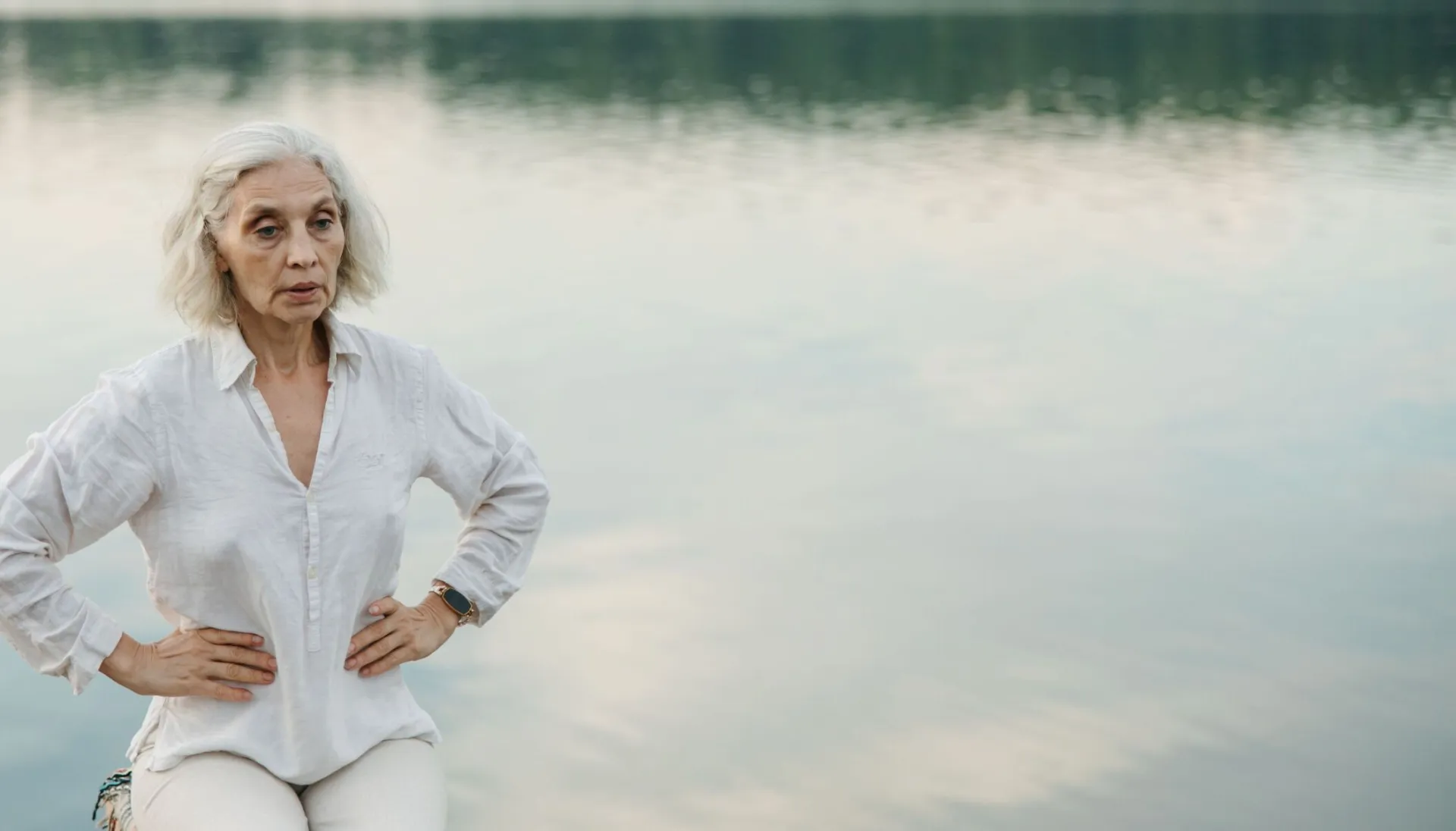 Elderly woman practicing mindfulness by a serene lake amid nature's tranquility.
