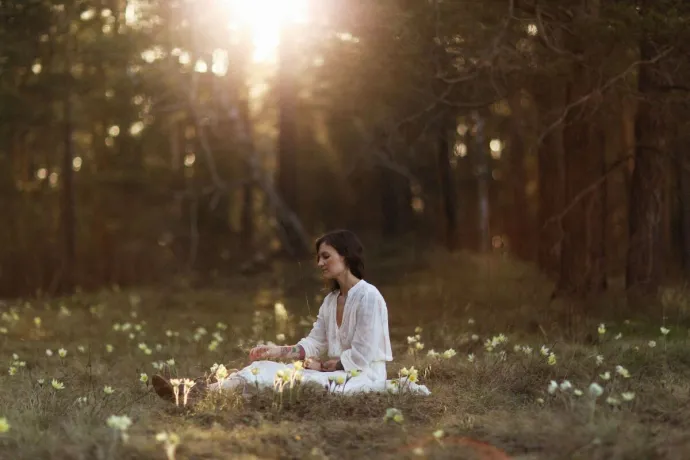 Woman meditates outdoors in a serene forest clearing with sunlight filtering through the trees.
