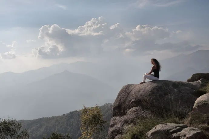 A woman enjoys serene solitude, sitting on a mountain rock with a vast view of the sunrise landscape.