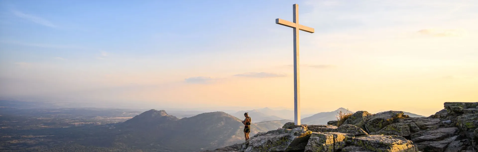 A hiker stands by a mountain cross at sunrise in Spain, surrounded by scenic vistas and serene skies.
