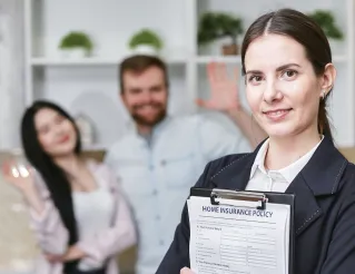 A smiling businesswoman holding a home insurance policy with clients in the background.