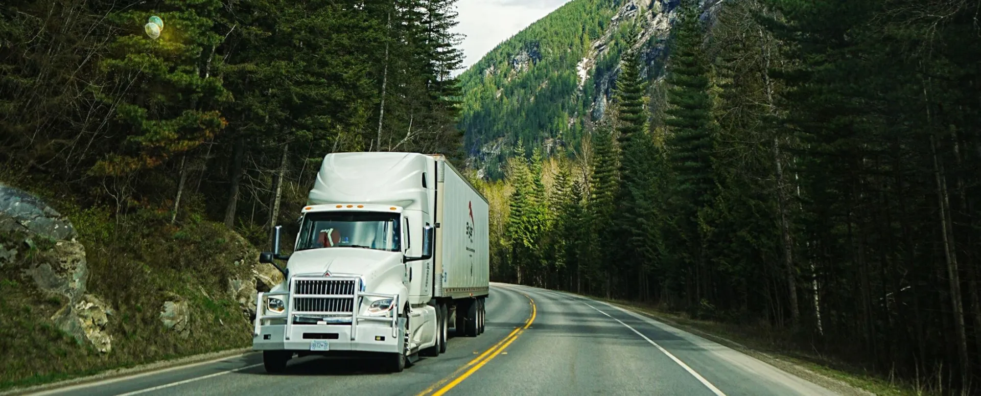 White delivery truck drives on a mountain road under a clear blue sky, surrounded by tall trees.