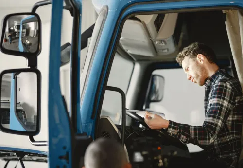 A male mechanic in a plaid shirt reads a checklist inside a truck cab, focused and serious.