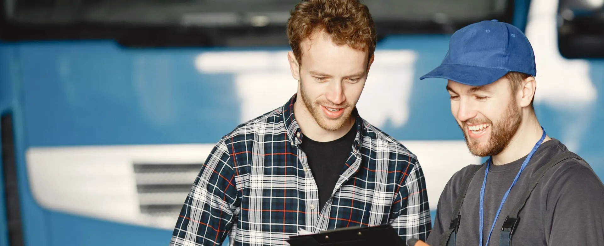 Two men discussing truck repairs in a garage: one in mechanic uniform, the other in a plaid shirt.