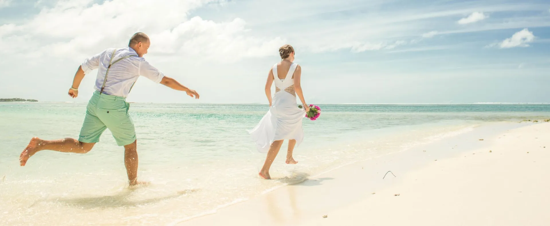 A couple in wedding attire joyfully running along a sunny tropical beach.