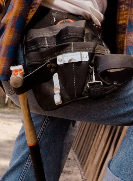 Close-up of a utility belt filled with tools on a person wearing jeans and a flannel shirt outdoors.