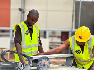 Two construction workers in high-visibility vests and helmets operating machinery at a building site.