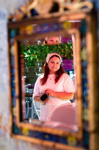 A woman holding a camera smiles into a colorful, decorative mirror, capturing her reflection in an outdoor setting.