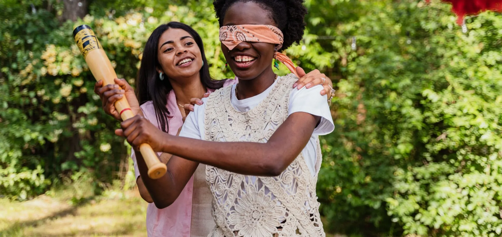 Two women laughing and playing a blindfolded game with a bat outdoors on a sunny day.