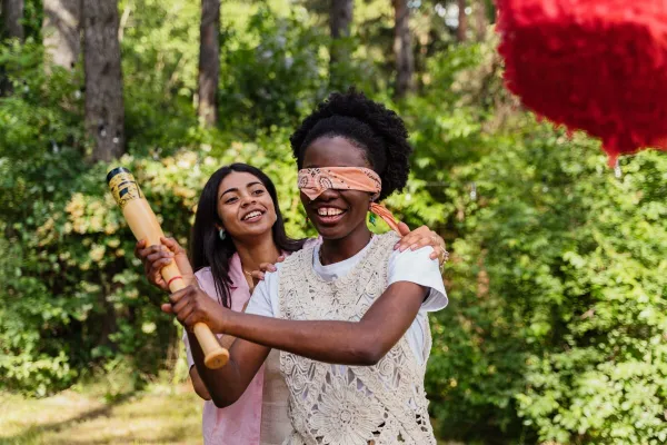 Two women laughing and playing a blindfolded game with a bat outdoors on a sunny day.