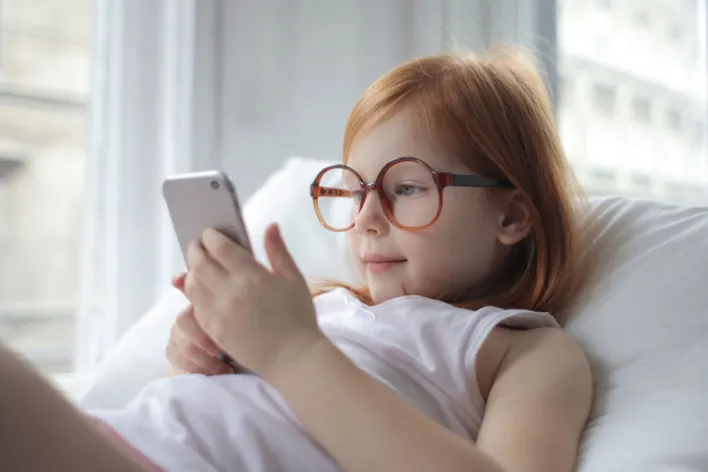 Adorable young girl with glasses engaged in a smartphone indoors, capturing a moment of leisure and technology.