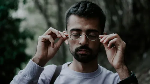 Close-up portrait of a man adjusting his eyeglasses in an outdoor setting, with a thoughtful expression.