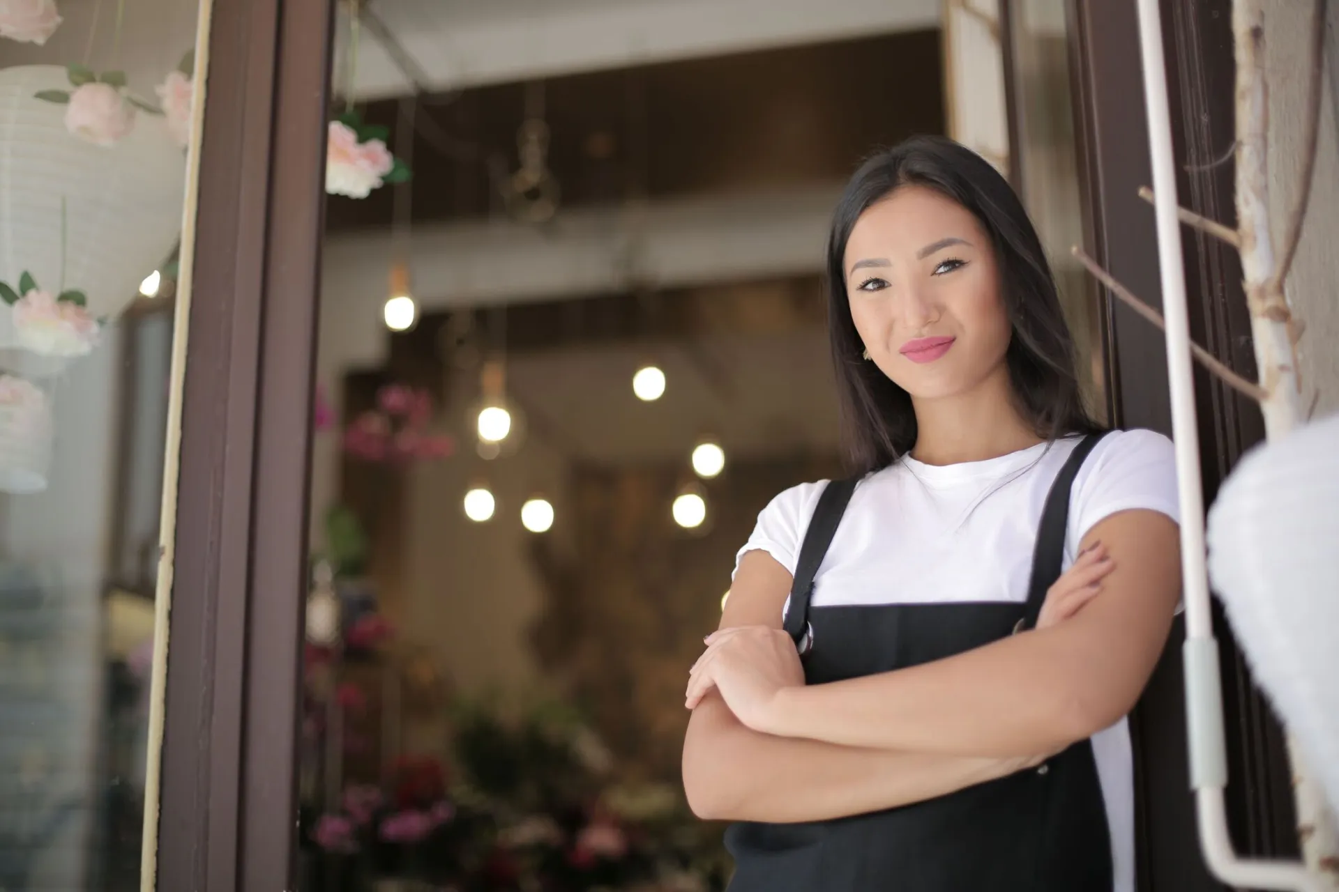From below of young confident Asian female standing with crossed arms at entrance of floral shop and smiling at camera