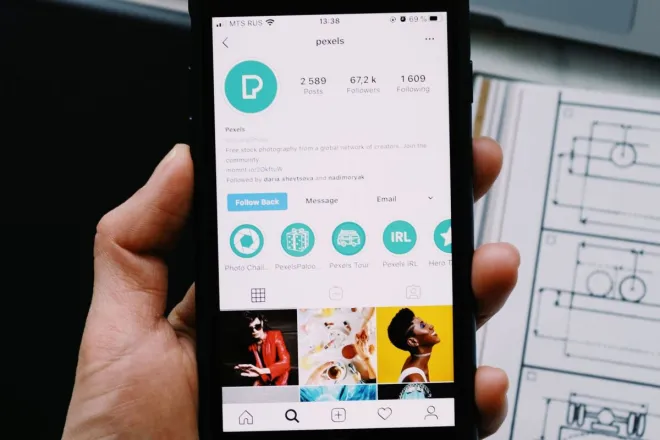 Hand holding smartphone displaying social media profile with tech gadgets on desk.