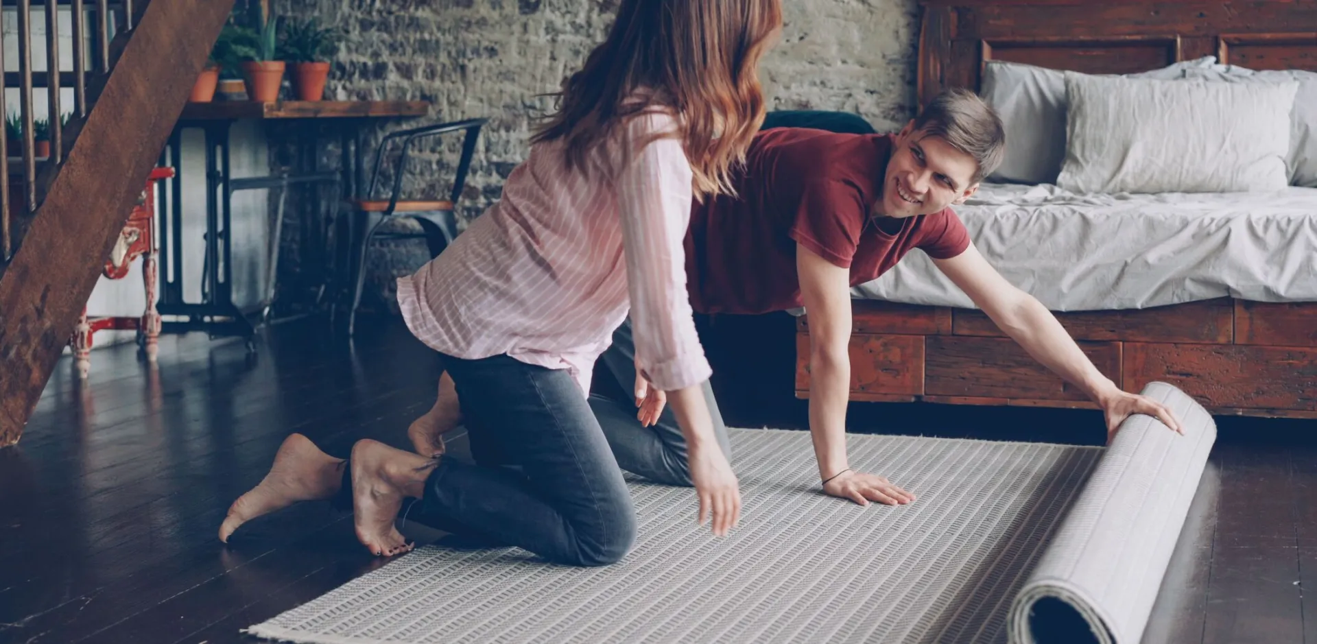 A couple unrolling a carpet in a stylish, industrial-themed bedroom with a brick wall.