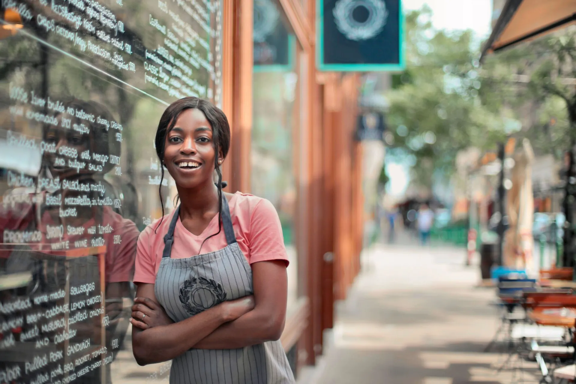 Smiling waitress in an apron standing outside a restaurant, displaying a welcoming atmosphere.