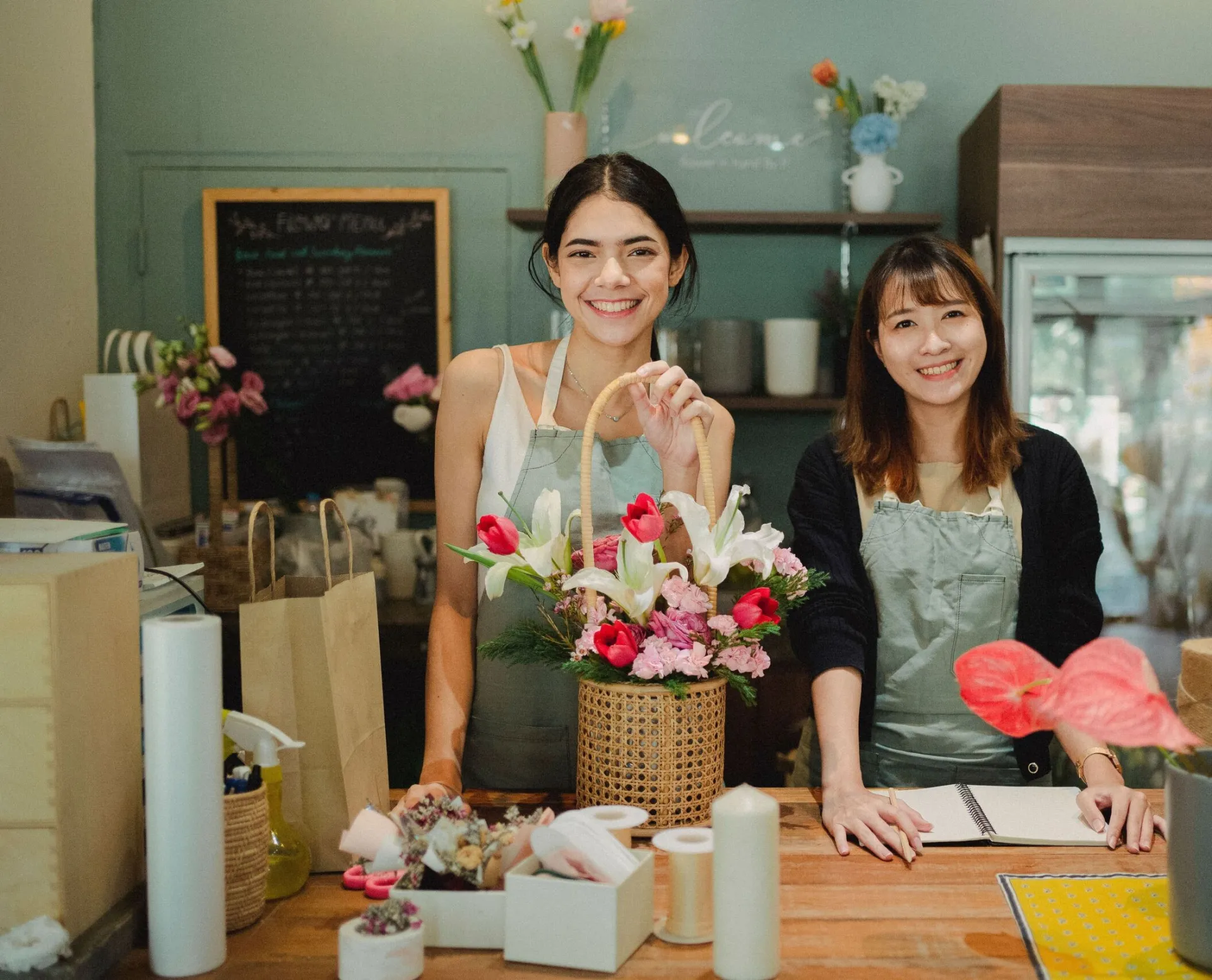 Positive diverse florists in casual clothes and aprons standing at counter with bouquets of flowers in cozy floristry store and looking at camera