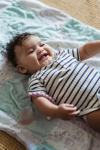 From above cute ethnic baby lying on linen on floor in light bedroom and looking away happily