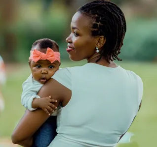 A mother holding her baby girl with a pink bow in a serene outdoor park setting.